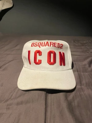 Vit Dsquared2 ICON keps - Snygg vit keps från Dsquared2 med röd brodyr där det står 'ICON' framtill och Dsquared2 både fram och bak. Klassisk böjd skärm och justerbart spänne bak. Tillverkad i bomull för en bekväm och cool look.