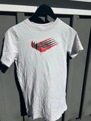 Vit Nike t-shirt med röd logga - Säljer en vit t-shirt från Nike med ett coolt tryck av flera swoosh-loggor i rött och svart på bröstet. Klassisk passform och rund halsringning. Perfekt för dig som gillar sportig och enkel stil.