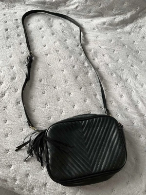 Svart axelväska med quiltad design - Snygg svart axelväska i konstläder med quiltat mönster i chevron-stil. Väskan har justerbar axelrem med silverfärgat spänne och dekorativ tofs på sidan. Perfekt storlek för det viktigaste och ger en trendig touch till din outfit.