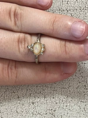 Guldfärgad ring med opal och stenar - Unik guldfärgad ring med en oval opal i mitten som skimrar i olika färger. Ringen har små klara stenar på sidorna av opalen som ger extra bling. Perfekt för dig som gillar smycken med lite vintagekänsla och vill sticka ut.