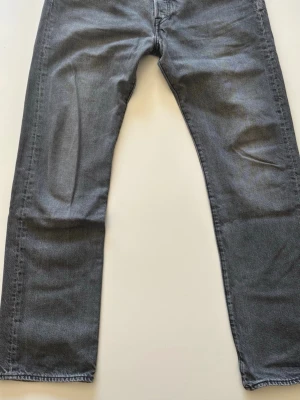 Levis 501 W34 L32 - Säljer ett par klassiska grå jeans från Levi's med rak passform och femficksmodell. Jeansen har en snygg tvättad look och är tillverkade i mjuk denim. Perfekta för dig som gillar en enkel och tidlös stil. STORLEK: W34 L32