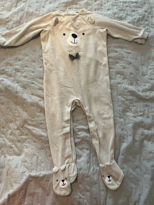 Beige body med björnansikte och öron - Supersöt beige body med långa ärmar och ben, dekorerad med ett broderat björnansikte, små öron på axlarna och en liten rosett. Fötterna har gulliga björnansikten. Knäppning längs benen för enkel påklädning. Mjuk och skön bomullsmix.