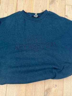 Mörkblå sweatshirt Keywest Bay - Mörkblå sweatshirt med broderad text 'ROAD TO KEYWEST BAY 1989' på bröstet. Tröjan har rund halsringning, ribbade muddar vid ärmslut och nederkant samt en avslappnad passform. Perfekt för en casual och stilren look.