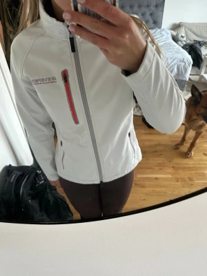 Vit softshelljacka från Everest - Snygg vit softshelljacka från Everest med hög krage och dragkedja framtill. Jackan har en röd detalj på bröstfickan och två sidofickor med dragkedja. Perfekt för dig som gillar sportig stil och funktionella plagg.