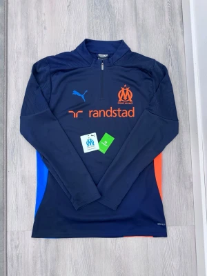 Marinblå Marseille träningströja Puma - Snygg marinblå långärmad träningströja från Puma med Olympique de Marseille-logga och orangea samt blå detaljer på sidorna. Halvlång dragkedja framtill och tryckta sponsorloggor. Tillverkad i funktionsmaterial som andas.