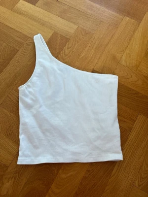 Vit ribbad oneshoulder topp - Säljer en vit ribbad oneshoulder topp i stretchigt material. Toppen har en asymmetrisk axel och är croppad, vilket ger en trendig och fräsch look. Perfekt att matcha med höga jeans eller kjol för en clean stil.