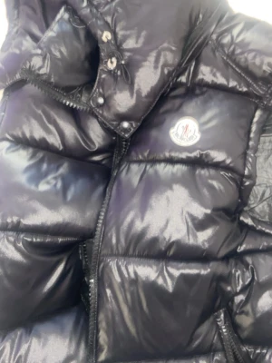 Svart dunväst från Moncler - Snygg svart dunväst från Moncler med glansig finish och klassisk logotyp på bröstet. Västen har hög krage, dragkedja och tryckknappar framtill. Perfekt för lager-på-lager och ger en trendig look till din outfit.