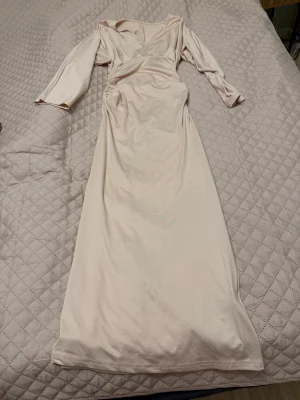 Ljus långklänning med v-ringning - Elegant ljus långklänning med v-ringad hals och långa ärmar. Klänningen har en smickrande omlott-detalj i midjan och är tillverkad i ett mjukt, följsamt material. Perfekt för speciella tillfällen.