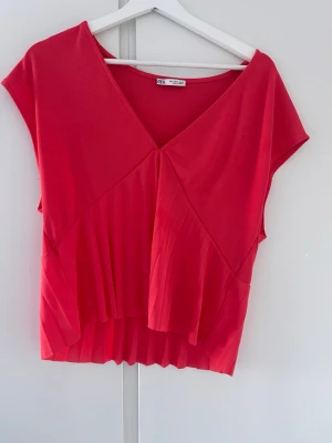 Röd/orange v-ringad topp från Zara - Säljer en snygg röd topp från Zara med v-ringning och plisserade detaljer framtill. Toppen är ärmlös och har en modern, croppad passform som ger en cool look. Perfekt att styla med jeans eller kjol för en trendig outfit.