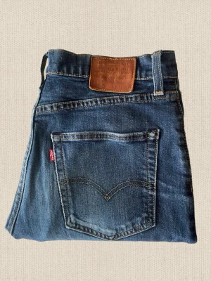 Levi's - Säljer ett par klassiska blå Levi's jeans i denim med femficksmodell och ikonisk röd etikett på bakfickan. Jeansen har en brun läderpatch i midjan och raka ben. Perfekta för en avslappnad och tidlös stil.