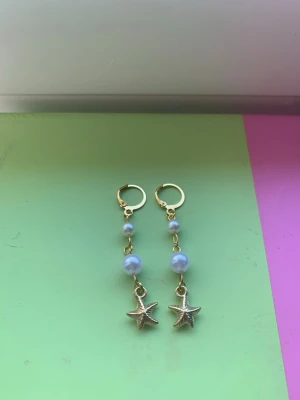 Guldörhängen med pärlor och sjöstjärnor - Säljer ett par trendiga örhängen i guldfärg med två ljuslila pärlor och en liten sjöstjärneberlock längst ner. Perfekta för dig som gillar havsinspirerade accessoarer och vill ha något unikt till din stil.
