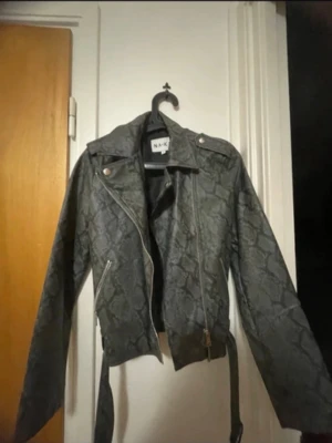 Grå/ svart ormmönstrad skinnjacka NAKD - Säljer en grå skinnjacka från NA-KD med coolt ormmönster. Jackan har klassisk bikerstil med sned dragkedja, axelklaffar och bälte i midjan. Silverfärgade detaljer och fickor med dragkedja ger extra edge till looken.