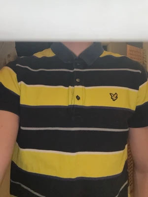  Lyle & Scott piké - Säljer en kortärmad pikétröja från Lyle & Scott med breda marinblå, gula och vita ränder. Tröjan har klassisk krage, knappslå med tre knappar och den ikoniska gula loggan broderad på bröstet. Tillverkad i mjuk bomull som känns skön mot huden.