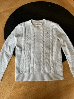 Ljusgrå kabelstickad tröja Hollister - Ljusgrå kabelstickad tröja från Hollister med rund halsringning och ribbade muddar. Tröjan har ett diskret Hollister-logotyp broderat på bröstet och är gjord i mjukt material. Klassisk kabelstickning framtill ger en snygg och tidlös look.