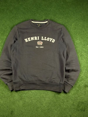 Mörkblå sweatshirt från Henri Lloyd - Mörkblå sweatshirt från Henri Lloyd med rund halsringning och broderad logga samt texten 'Henri Lloyd Est. 1963' på bröstet. Tröjan har långa ärmar och ribbade muddar vid ärmslut och nederkant. Perfekt för en avslappnad och stilren look.