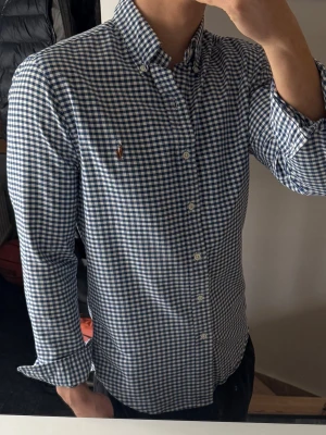 Rutig blå skjorta från Polo Ralph Lauren - Klassisk rutig skjorta från Polo Ralph Lauren i blått och vitt. Skjortan har lång ärm, knappar framtill och button-down krage. Tillverkad i mjuk bomull och har den ikoniska broderade loggan på bröstet. Perfekt för en clean och stilren look.