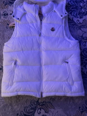Vit dunväst från Moncler - Snygg vit dunväst från Moncler med hög krage och avtagbar huva. Västen har två dragkedjeförsedda fickor fram och en Moncler-logga på bröstet. Perfekt för lager på lager och riktigt trendig look.