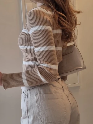 Beige och vit randig stickad tröja NA-KD - Stickad tröja från NA-KD i beige med vita horisontella ränder. Tröjan är ribbstickad, har en figurnära passform och långa ärmar. Perfekt att matcha med jeans eller kjol för en snygg look.