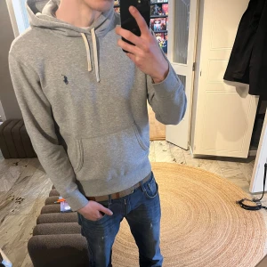 Ralph Lauren hoodie - Snygg grå hoodie från Polo Ralph Lauren med klassisk huva och vita dragsnören. Tröjan har en liten mörkblå logga broderad på bröstet och en stor magficka framtill. Tillverkad i mjukt bomullsmaterial som är skönt att bära.