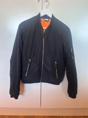 Svart bomberjacka med orange foder - Klassisk svart bomberjacka med orange foder. Jackan har ribbade muddar, två snedställda fickor med dragkedja och en ärmficka med dragkedja. Säljer den för den kommer inte till användning och är lite kort i armarna på mig 