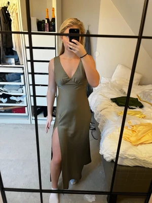 Olivgrön långklänning med slits - Säljer en olivgrön långklänning med djup v-ringning och hög slits på ena sidan. Klänningen är ärmlös och har en figurnära passform som framhäver siluetten. Perfekt för dig som vill ha en stilren och fin klänning. Passar perfekt till bal och bröllop💞