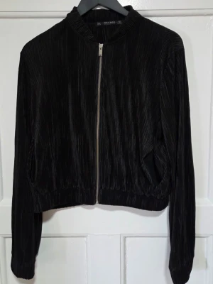 Svart ribbad plisserad tröja Zara - Snygg svart tröja från Zara med plisserat tyg och dragkedja framtill, samt två fickor. Tröjan har en kort, boxig passform, ribbade muddar och rundad krage. Perfekt för dig som vill ha en trendig och stilren look.