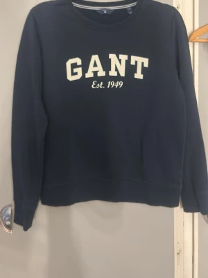 Mörkblå sweatshirt från GANT - Mörkblå sweatshirt från GANT med vit text 'GANT Est. 1949' på bröstet. Klassisk rund halsringning och långa ärmar. Tillverkad i mjukt material som passar perfekt till en avslappnad stil.
