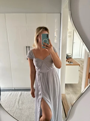 Ljuslila långklänning med slits - Säljer en  ljuslila långklänning med v-ringad byst, korta ärmar och en snygg slits framtill från Nelly. Klänningen har draperad detalj över bysten. Kan användas som sommarklänning eller balklänning. 