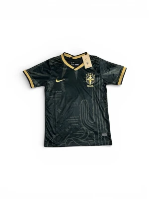 Svart Brasilien Nike fotbollströja☀️ - Snygg svart matchtröja från Nike med Brasilien-emblem och guldiga detaljer. Tröjan har korta ärmar, V-ringning och diskret mönster i tyget. Tillverkad i lätt och ventilerande material, perfekt för fotboll eller träning.