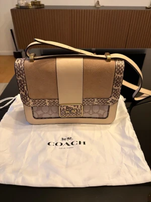 Coach beige axelväska med mönster - Snygg axelväska från Coach i beige och ljusbruna toner med ormmönstrade detaljer och klassiskt Coach-mönster. Väskan har guldfärgade metalldetaljer, justerbar axelrem och en rektangulär form. Perfekt för dig som vill ha en trendig och stilren accessoar.