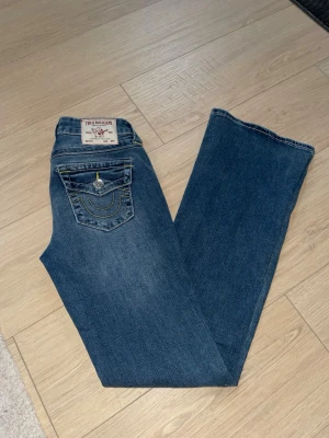 Blå True Religion jeans med fickdetaljer - Säljer ett par blå jeans från True Religion. ikoniska fickor bak med gul kontrastsöm och silverknappar. Klassisk patch i midjan och snygg tvätt. Perfekta för dig som gillar streetwear och vill ha något som sticker ut. Nyskick!! Nypris är 1200kr storlek W25