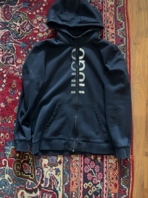 Mörkblå hoodie med dragkedja - Snygg mörkblå hoodie från Hugo med dragkedja framtill och stor vit logga vertikalt på bröstet. Tröjan har huva med snörning och en stor magficka. Tillverkad i mjukt sweatshirtmaterial och har ribbade muddar vid ärmar och nederkant.