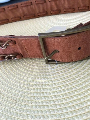 Brunt bälte med kedjedetaljer - Snyggt brunt bälte  med metallkedjor längs kanterna och en robust metallspänne. Perfekt accessoar för att lyfta din outfit med en cool och unik touch. Passar till jeans eller kjol för en trendig look.