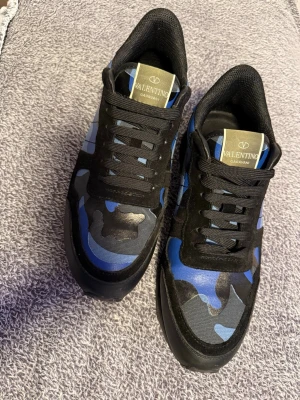 Valentino blå camo sneakers - Snygga sneakers från Valentino med blått och svart camo-mönster. Skorna har svarta snören, svart sula och detaljer i mocka och textil. Märkeslogga på plösen och undersidan av sulan. Perfekta för dig som vill sticka ut med en unik stil.