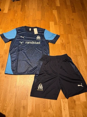 Marseille summer set Navy blue  - Säljer ett mörkblått träningsset från Olympique de Marseille och Puma. Setet består av en t-shirt med ljusblå detaljer och klubbmärke samt matchande shorts med klubbmärke och Puma-logga. Materialet är lätt och andas, perfekt för fotbollsträning.
