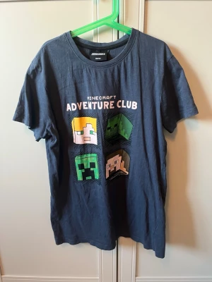 Minecraft Adventure Club t-shirt - Mörkblå t-shirt från Minecraft med trycket 'Adventure Club' och fyra klassiska Minecraft-karaktärer på framsidan. T-shirten har rund hals och korta ärmar, perfekt för alla som älskar gaming och vill ha en avslappnad stil.