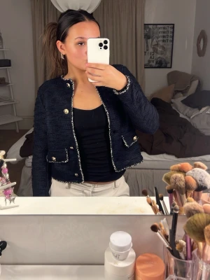 Mörkblå tweedkofta med gulddetaljer - Snygg mörkblå tweedkofta med guldfärgade knappar och dekorativa fickor framtill. Koftan har vit- och svartmönstrade kanter och är kort i modellen. Perfekt att styla över ett linne eller t-shirt för en chic look. Köpt på Sellpy 