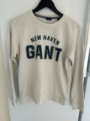 Vit sweatshirt från GANT - Snygg vit sweatshirt från GANT med trycket 'NEW HAVEN GANT' i svart på bröstet. Tröjan har rund halsringning och långa ärmar. Perfekt för en avslappnad och sportig stil.