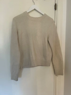 Beige stickad tröja med rund hals - En enkel och stilren beige stickad tröja med rund halsringning och ribbade muddar vid ärmslut och nederkant. Tröjan har en rak passform och är perfekt för lager på lager. Materialet ser mjukt och bekvämt ut, perfekt för kyligare dagar.