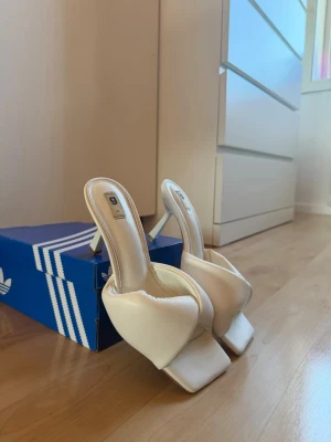 Vita quiltade slip-in pumps - Säljer ett par trendiga vita pumps med quiltad bred rem och öppen häl. Skorna har smal klack och fyrkantig tå, vilket ger en modern look. Perfekta för dig som vill sticka ut med stilrena och bekväma slip-ins i syntetmaterial. Använd en gång på ett kalas för en vän och inte mer än så, helt nya men har inte användning av dom längre då jag har för mycket skor. 