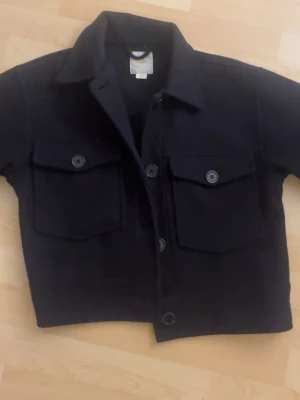 Svart overshirt med fickor - Svart overshirt med två stora bröstfickor och knappar framtill. Klassisk krage och raka ärmar. Använd sparsamt. 