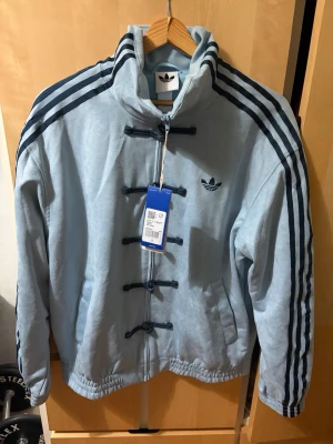 Adidas chinese lunar jacka - Ljusblå jacka från adidas med klassiska mörkblå ränder på ärmarna och adidas-logga på bröstet. Jackan har unik snörning framtill istället för dragkedja och är tillverkad i ett mjukt material. Perfekt för dig som vill sticka ut med sportig stil.