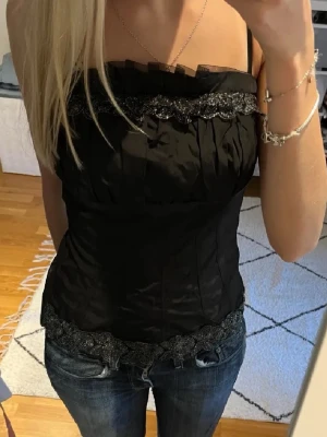 Svart korsettopp med spetsdetaljer - Snygg svart korsettopp med glittriga spetsdetaljer upptill och nedtill. Axelbandsmodell med figurnära passform och lätt glansigt material. Perfekt för dig som vill ha en trendig och elegant look.