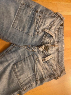 Ljusblå bootcut jeans med slitning från Lee  - Säljer ett par ljusblå jeans med bootcut-modell och stora framfickor. Jeansen har en tydlig slitning på vänster lår och coola detaljer som dubbla bälteshällor. Perfekta för en avslappnad och trendig look.