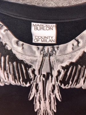 Marcelo Burlon gråvit - Säljer en svart sweatshirt från Marcelo Burlon County of Milan med ikoniskt vitt och grått vingmönster runt halsen. Tröjan har rund halsringning och ribbade muddar vid ärmslut och nederkant. Perfekt för dig som gillar streetwear och unik design.