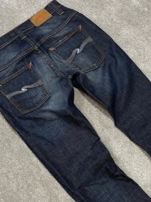 Mörkblå Nudie Jeans - Säljer ett par mörkblå jeans från Nudie Jeans med klassisk femficksdesign och kontrastsömmar. Jeansen har raka ben och detaljerade bakfickor med Nudie Jeans signatursöm. Tillverkade i slitstark denim, perfekta för en avslappnad stil.