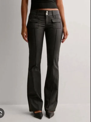 Nelly Coated jeans  - Snygga svarta jeans med utsvängda ben och markerade sömmar framtill. Jeansen har två stora fickor framtill och stängs med två vita knappar. Perfekta för en trendig och cool look.