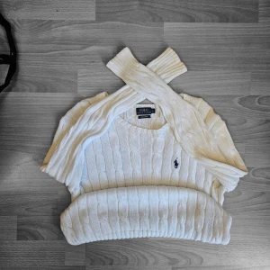 Vit kabelstickad tröja Polo Ralph Lauren - Klassisk vit cable knit tröja från Polo Ralph Lauren med fin flätstickning över hela, ribbad nederkant och ärmar samt det ikoniska broderade ponylogot på bröstet. Tillverkad i mjuk Pima Cotton. Passform: Damstorlek – sitter som en normal M (bra och vanlig passform, inte för tight eller oversized). Mått (platt): Axelbredd: ca 48–50 cm Pit to pit: ca 51–53 cm Längd: ca 66–68 cm Skick: Mycket bra skick – har använts ett fåtal gånger och är fortfarande i fint skick. Inga problem eller defekter. Pack