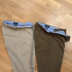 Två par chinos från Polo Ralph Lauren - Säljer två par chinos från Polo Ralph Lauren, ett par i beige och ett par i mörkbrunt. Klassisk rak passform med bälteshällor, sidofickor och knappstängning. Tillverkade i mjukt bomullstyg med blå innerfoderkant. Perfekta för en stilren och avslappnad look. Inte använda!!.    Storlek 31/32 på båda!