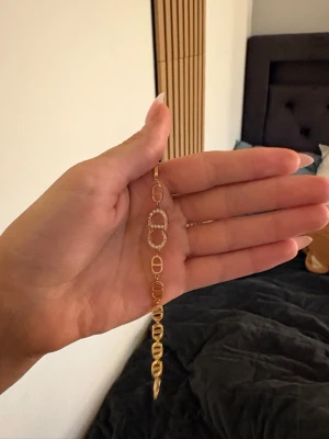 Guldigt armband med länkar och stenar - Snyggt armband i guldfärgad metall med ovala länkar och en större länk dekorerad med glittrande stenar. Armbandet har en modern design och passar perfekt till dig som gillar trendiga accessoarer.